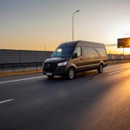 TRANSPORTER Piotr Kałęcki - Brązowy bus Mercedes-Benz Sprinter jadący autostradą o zachodzie słońca, widoczne światła w tle.