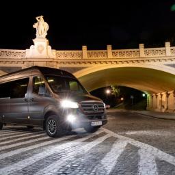TRANSPORTER Piotr Kałęcki - Szary Mercedes Sprinter na tle oświetlonego nocą mostu z rzeźbą na szczycie, zaparkowany na brukowanej drodze z namalowanymi pasami.