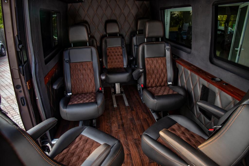Wnętrze luksusowego busa z czarno-brązowymi skórzanymi fotelami z diamentowym przeszyciem, drewnianą podłogą i oknami.