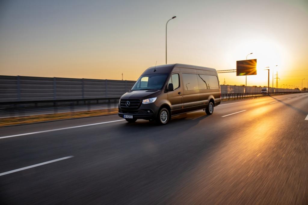 Brązowy bus Mercedes-Benz Sprinter jadący autostradą o zachodzie słońca, widoczne światła w tle.