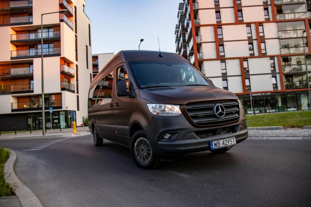 Brązowy van Mercedes-Benz Sprinter na tle nowoczesnych budynków mieszkalnych, z widocznym logo i tablicą rejestracyjną.