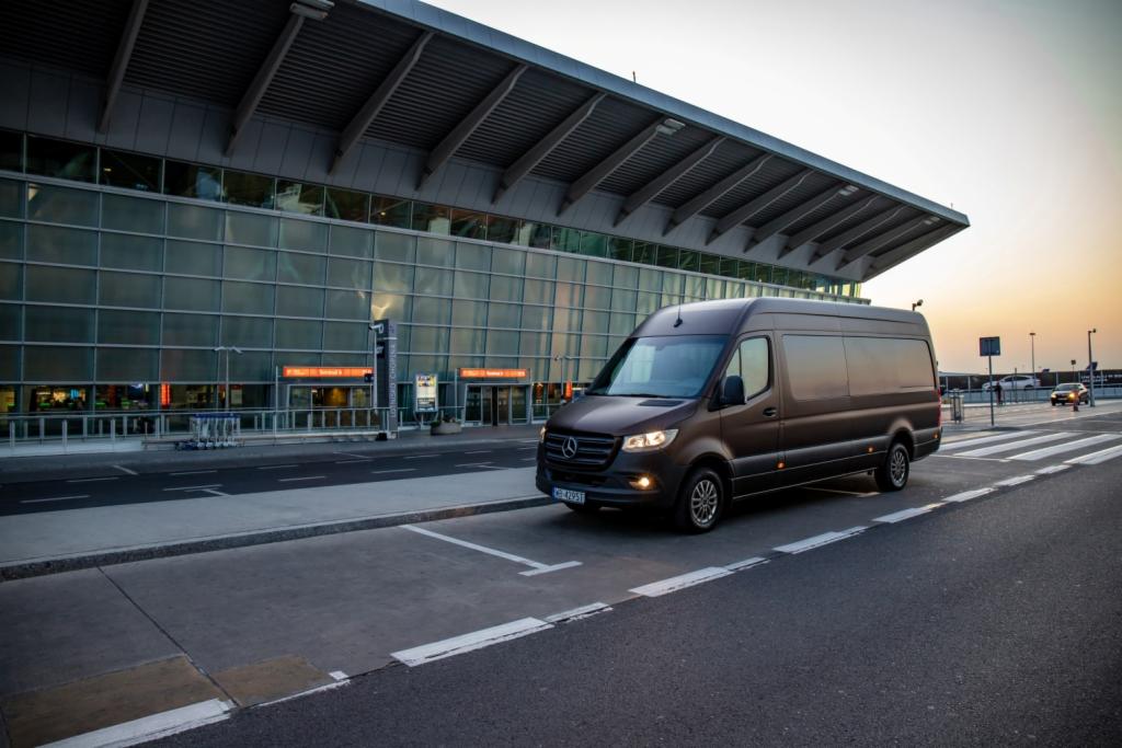 Brązowy van Mercedes-Benz Sprinter zaparkowany przed nowoczesnym budynkiem lotniska o szklanej fasadzie, z widocznymi w tle pasami dla pieszych i zachodzącym słońcem.