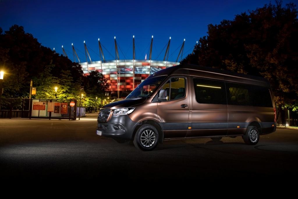 Brązowy bus na tle oświetlonego Stadionu Narodowego nocą, widok z boku.