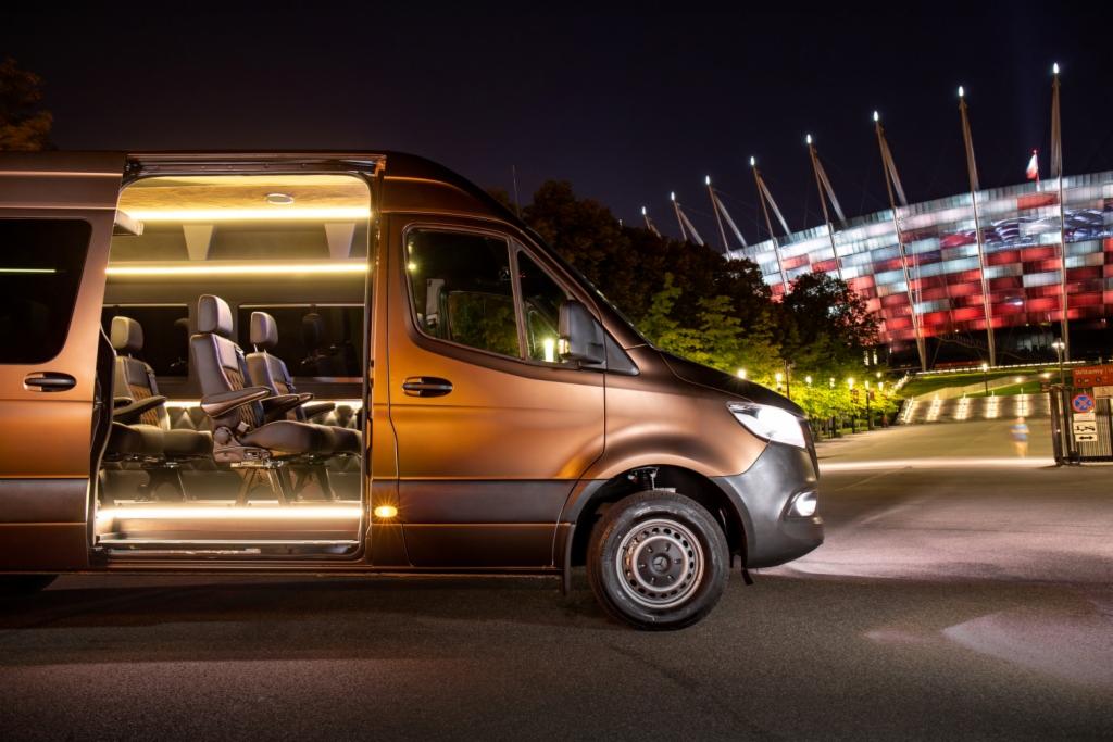 Brązowy van z otwartymi drzwiami, podświetlonym wnętrzem i luksusowymi fotelami na tle nocnego krajobrazu miejskiego ze stadionem.