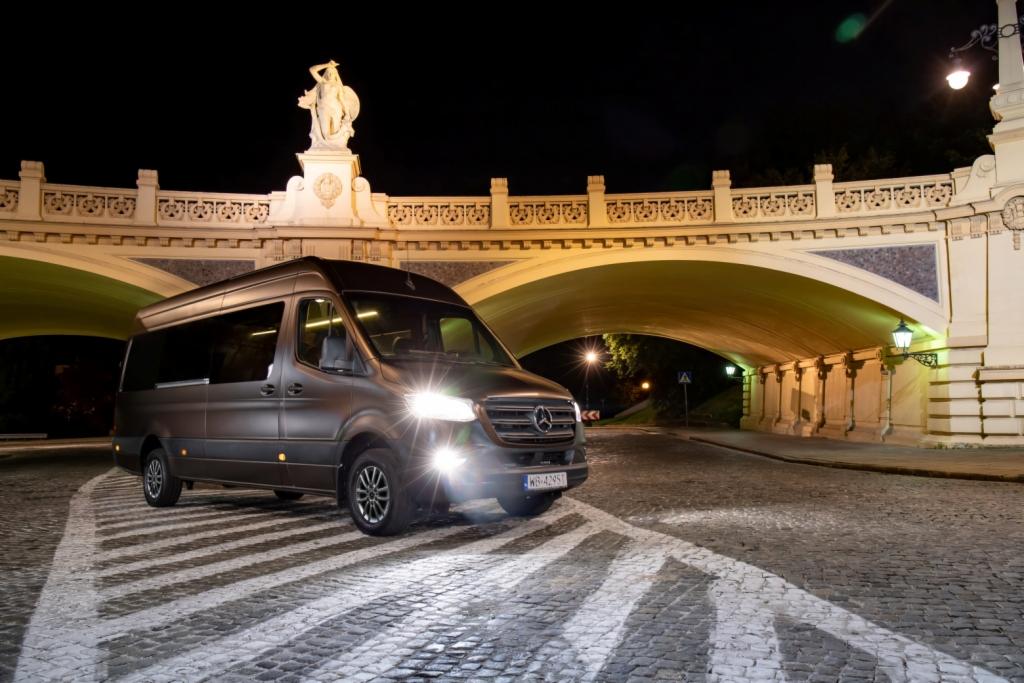 Szary Mercedes Sprinter na tle oświetlonego nocą mostu z rzeźbą na szczycie, zaparkowany na brukowanej drodze z namalowanymi pasami.