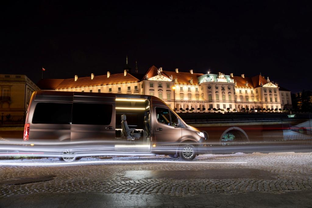 Luksusowy, brązowy van z otwartymi drzwiami na tle nocnego krajobrazu Warszawy, z widocznym Zamkiem Królewskim i smugami świateł.