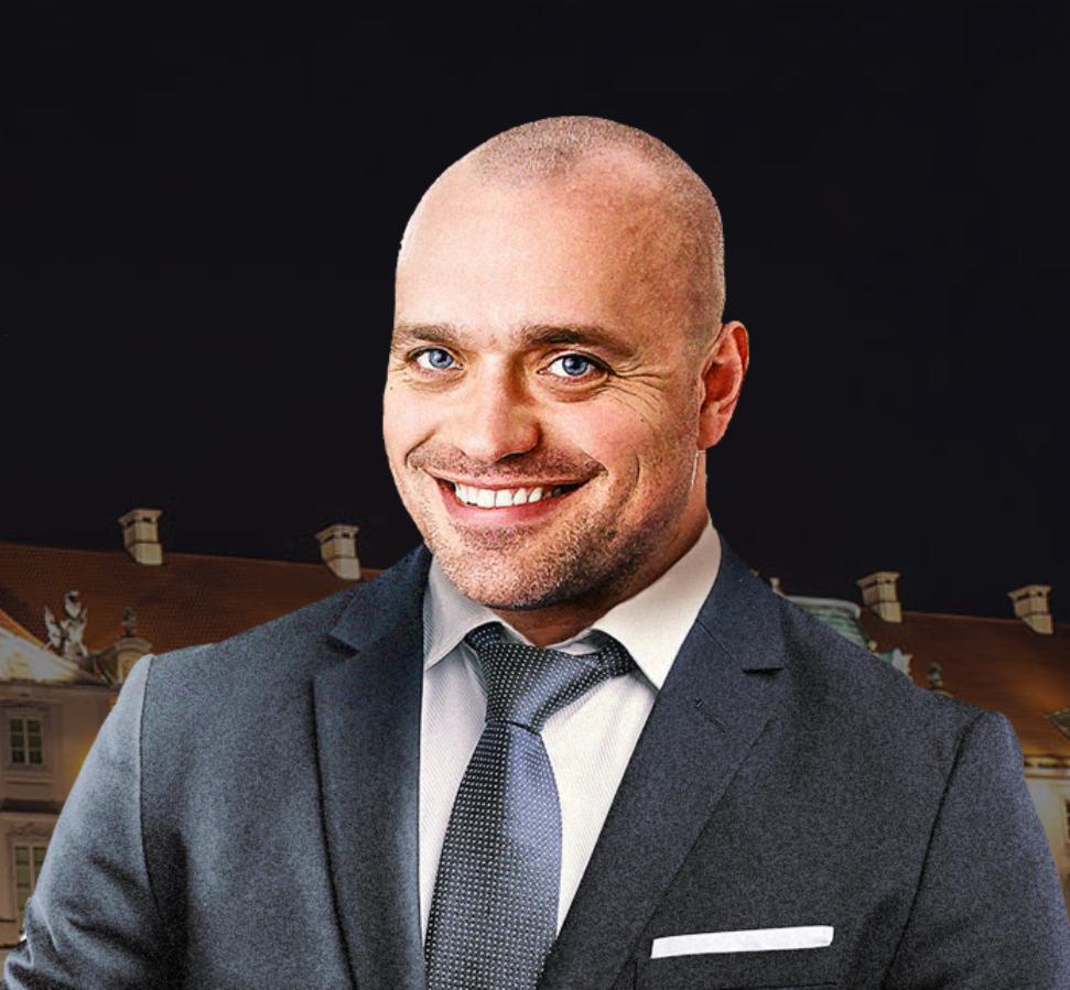 Portret uśmiechniętego mężczyzny w garniturze na tle budynku z czerwonym dachem i czarnym niebem.