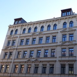 BUDMAR Producent okien i drzwi z drewna - Elewacja kamienicy z regularnym układem okien w czarnych ramach, widoczne detale architektoniczne i refleksy w szybach.