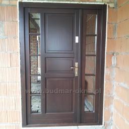 BUDMAR Producent okien i drzwi z drewna - Ciemnobrązowe drzwi zewnętrzne z szybami, zamontowane w surowym murze z cegły, widoczne przewody elektryczne na ścianie.