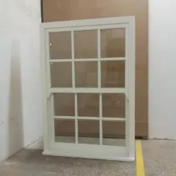 Białe, pionowe okno skrzynkowe z wieloma szybami, stojące na betonowej podłodze w magazynie.