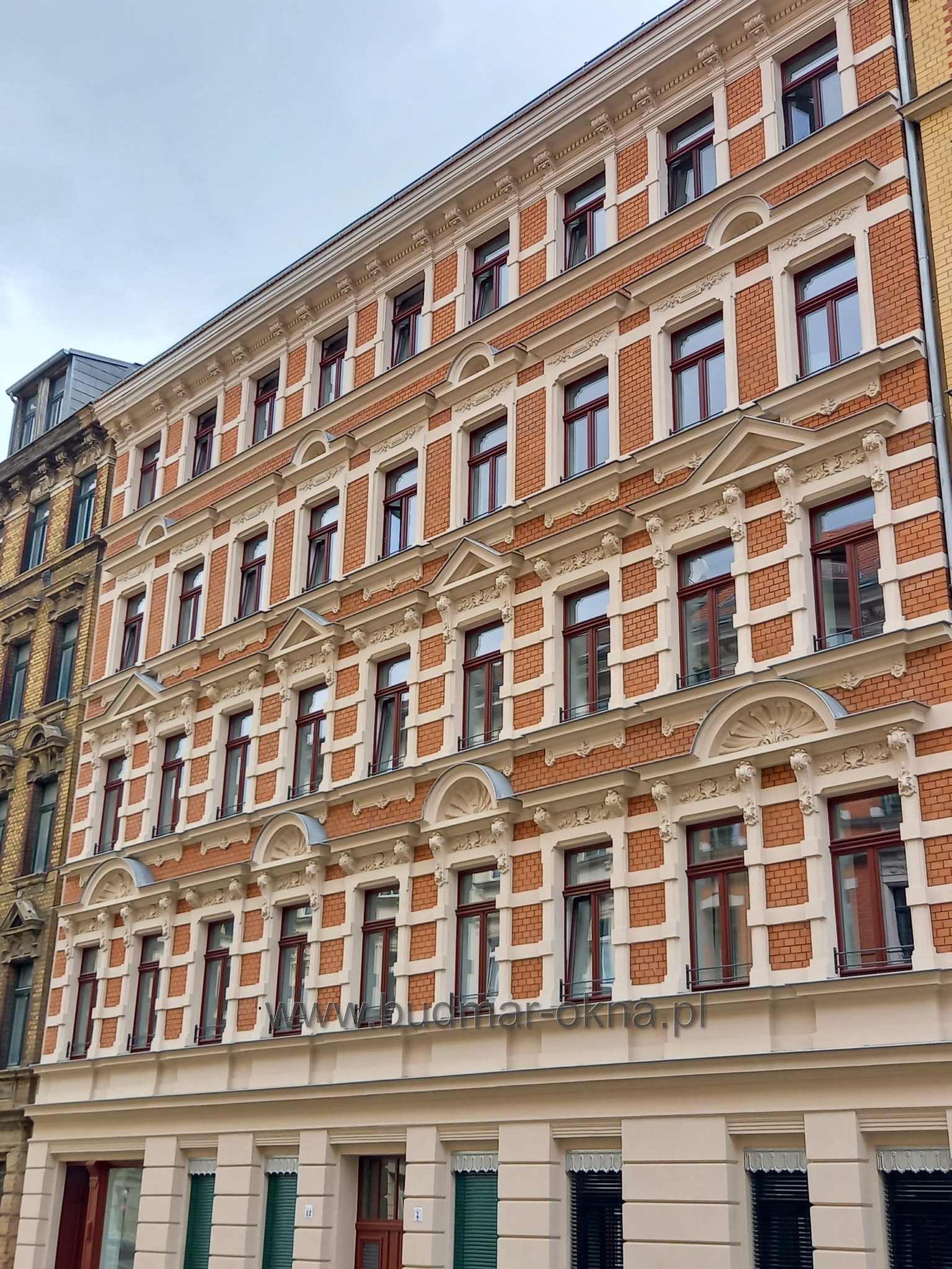 Fasada kamienicy z czerwonej cegły z rzędami okien z ciemną ramą, ozdobne gzymsy i elementy dekoracyjne w jasnym kremowym kolorze, widok z dołu ku górze.