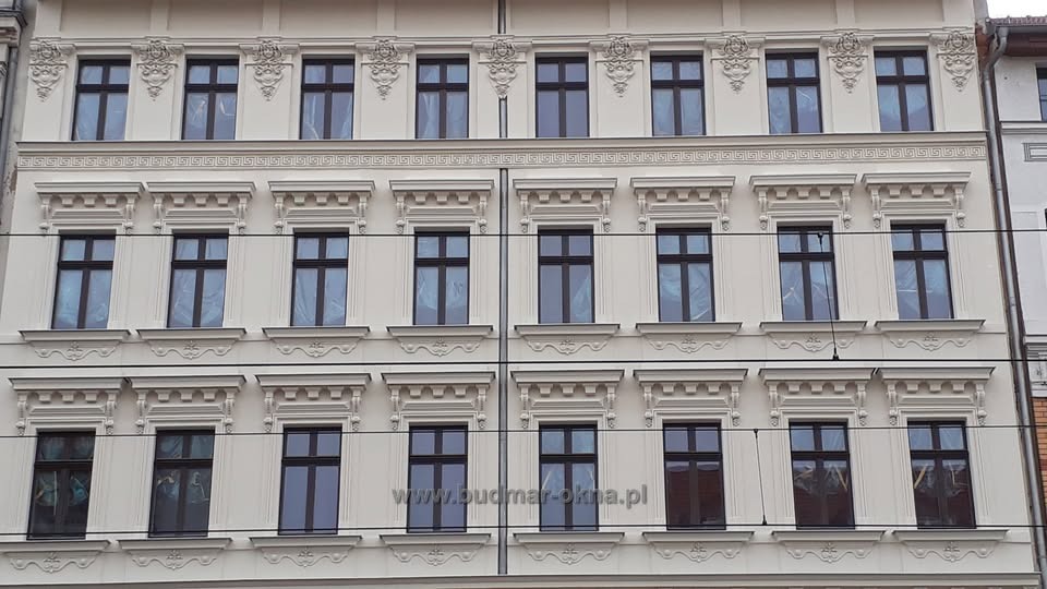 Fasada kamienicy po renowacji z nowymi oknami w ciemnych ramach, niektóre okna zabezpieczone folią ochronną, widoczny adres strony internetowej na elewacji.