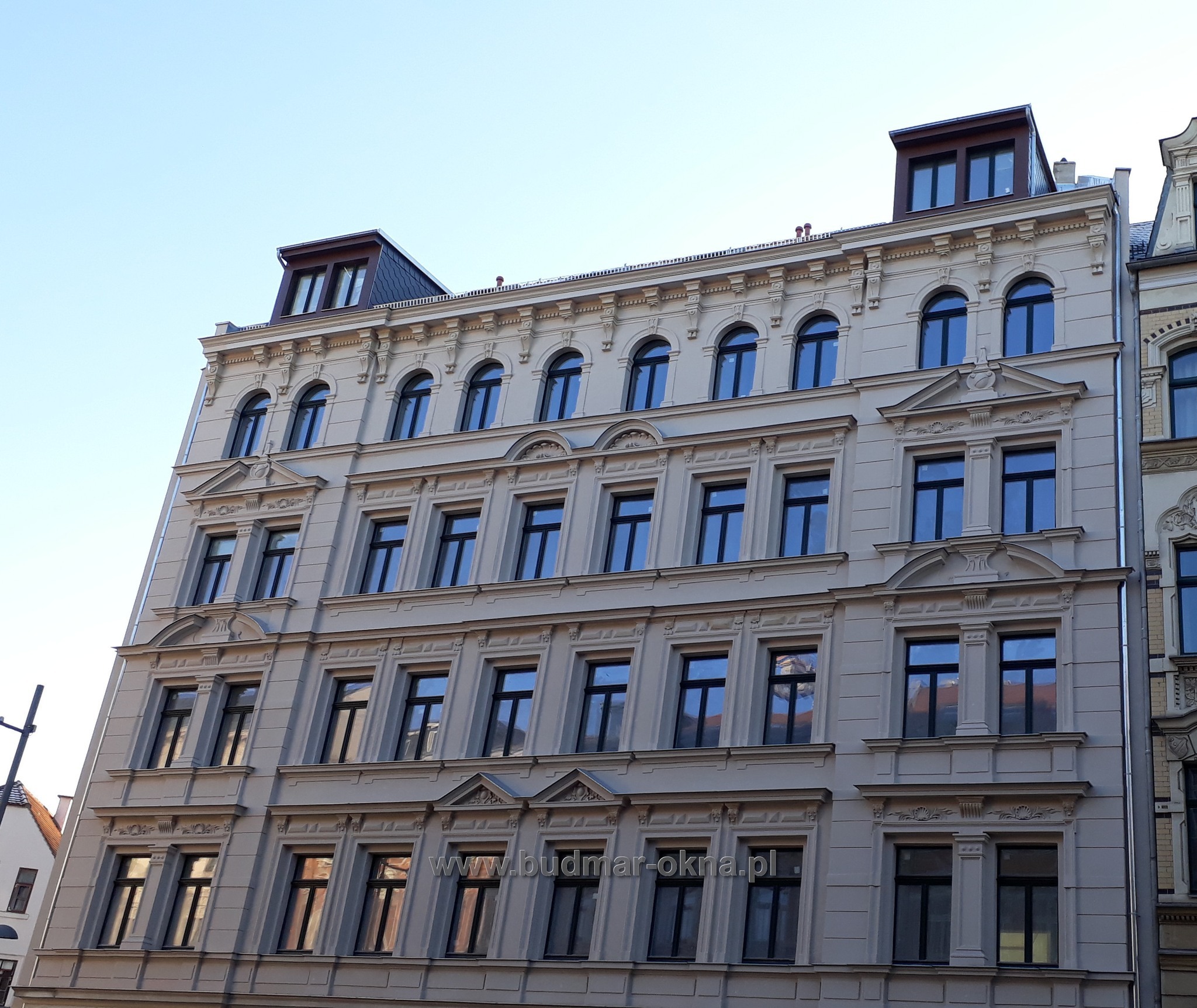 Elewacja kamienicy z regularnym układem okien w czarnych ramach, widoczne detale architektoniczne i refleksy w szybach.