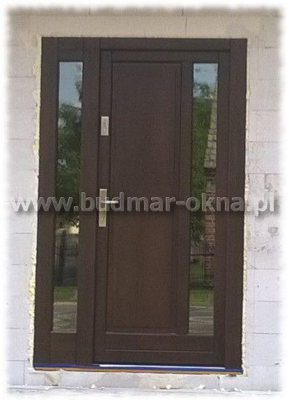 Brązowe drzwi wejściowe z dwoma bocznymi przeszkleniami, widoczny zamek i klamka, zamontowane w surowym murze z betonu komórkowego, z widocznym adresem strony internetowej na zdjęciu.