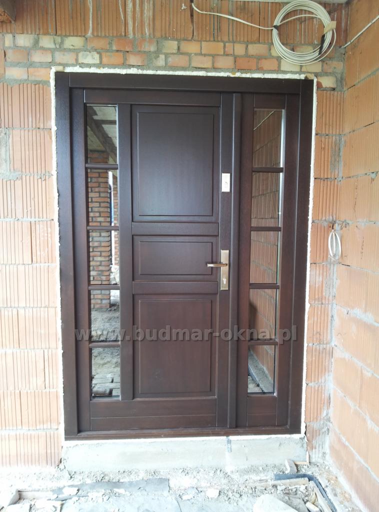 Ciemnobrązowe drzwi zewnętrzne z szybami, zamontowane w surowym murze z cegły, widoczne przewody elektryczne na ścianie.