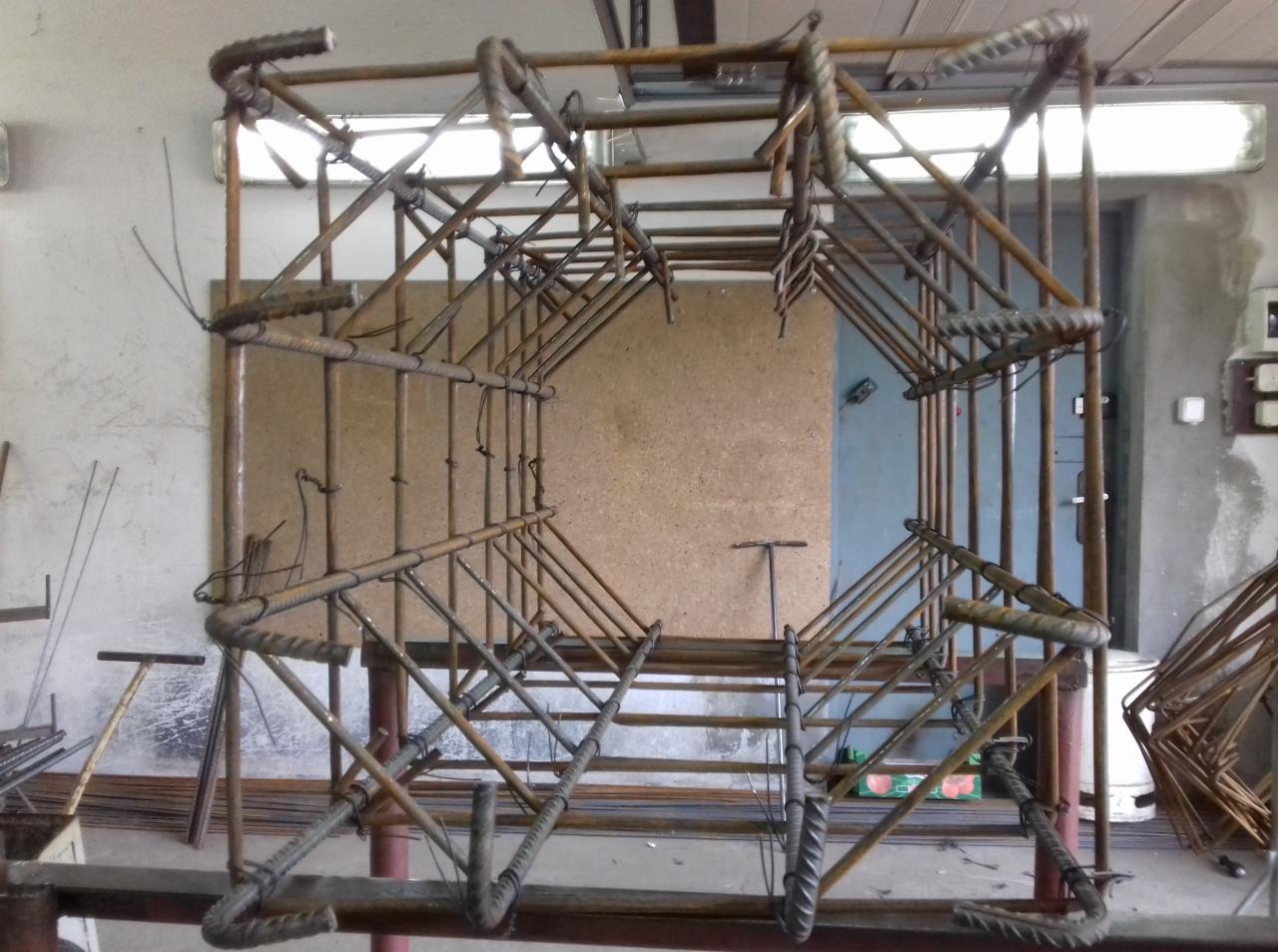 Szkielet z żebrowanych prętów zbrojeniowych, formujący przestrzenną konstrukcję o kształcie prostopadłościanu, przygotowany do zalania betonem, widoczny w surowym wnętrzu warsztatu.