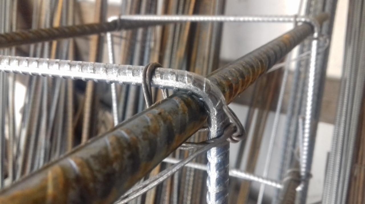 Połączenie dwóch prętów żebrowanych drutem wiązałkowym, widoczne ślady korozji na jednym z prętów, tło stanowią pionowo ułożone pręty.