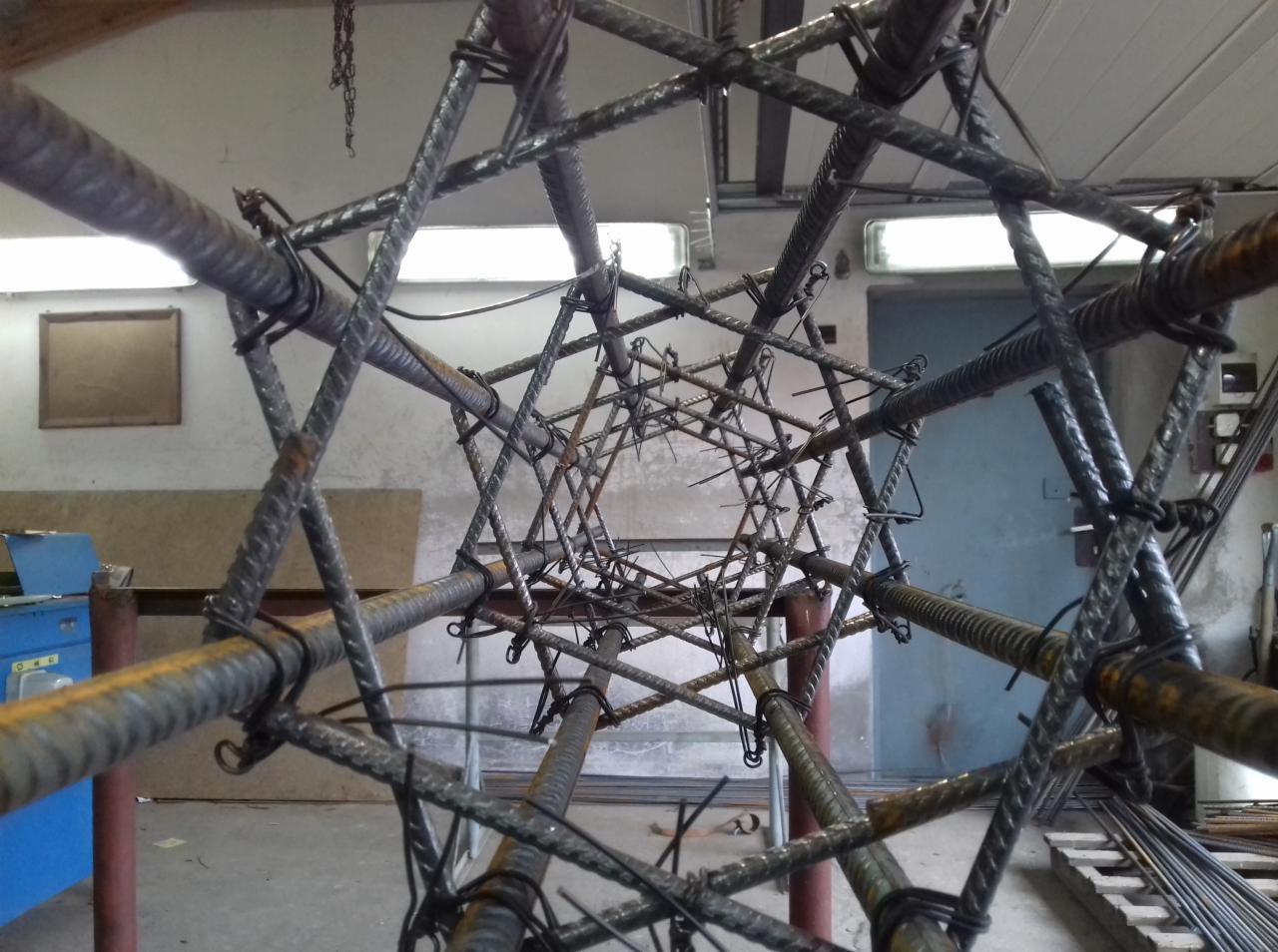 Szkielet zbrojenia kolumny z żebrowanej stali, widok od dołu, wiązany drutem, surowe wnętrze warsztatu w tle.