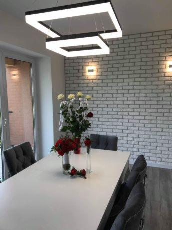 Jasne wnętrze jadalni z białym stołem, szarymi krzesłami i dekoracyjnymi kwiatami na tle ściany z białej cegły. Nowoczesna lampa sufitowa w kształcie dwóch kwadratów z oświetleniem LED.