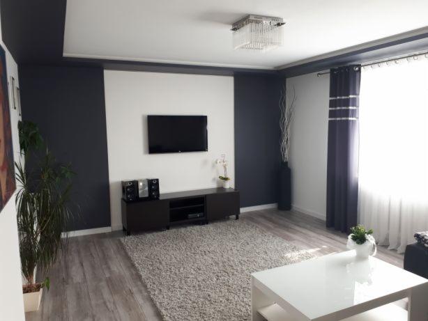 Salon po remoncie w odcieniach szarości i bieli z drewnianą podłogą, szarym dywanem, czarną ścianą i białym sufitem podwieszanym z nowoczesnym oświetleniem.