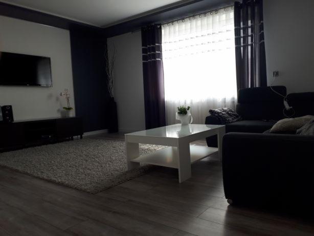 Nowoczesny salon z ciemną ścianą akcentującą, szarą wykładziną dywanową, białym stolikiem kawowym i ciemnymi meblami, oświetlony naturalnym światłem wpadającym przez okno z białymi firanami...