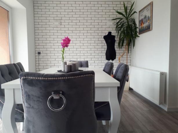 Jadalnia z białym stołem, szarymi welurowymi krzesłami z ozdobnymi pierścieniami, białą ceglaną ścianą, kwiatem w wazonie i manekinem krawieckim w tle.