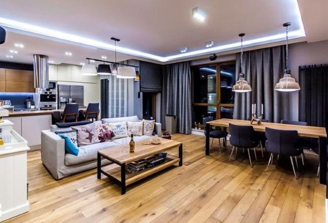 Przestronny salon z drewnianą podłogą, sofą z dekoracyjnymi poduszkami, stołem jadalnym z czarnymi krzesłami i otwartą kuchnią w tle, oświetlenie sufitowe LED.