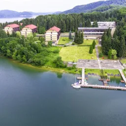 Widok z lotu ptaka na ośrodek wypoczynkowy nad jeziorem z przystanią dla żaglówek i budynkami z czerwonymi dachami otoczonymi lasem.