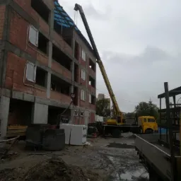 Żółty dźwig samochodowy podnosi materiały budowlane na budowę czteropiętrowego budynku z czerwonej cegły w pochmurny dzień.