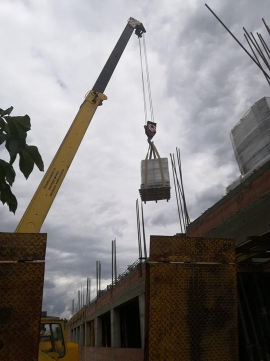Żółty dźwig podnosi paletę z materiałami budowlanymi na placu budowy, widoczne zbrojenia i fragment budynku w surowym stanie.