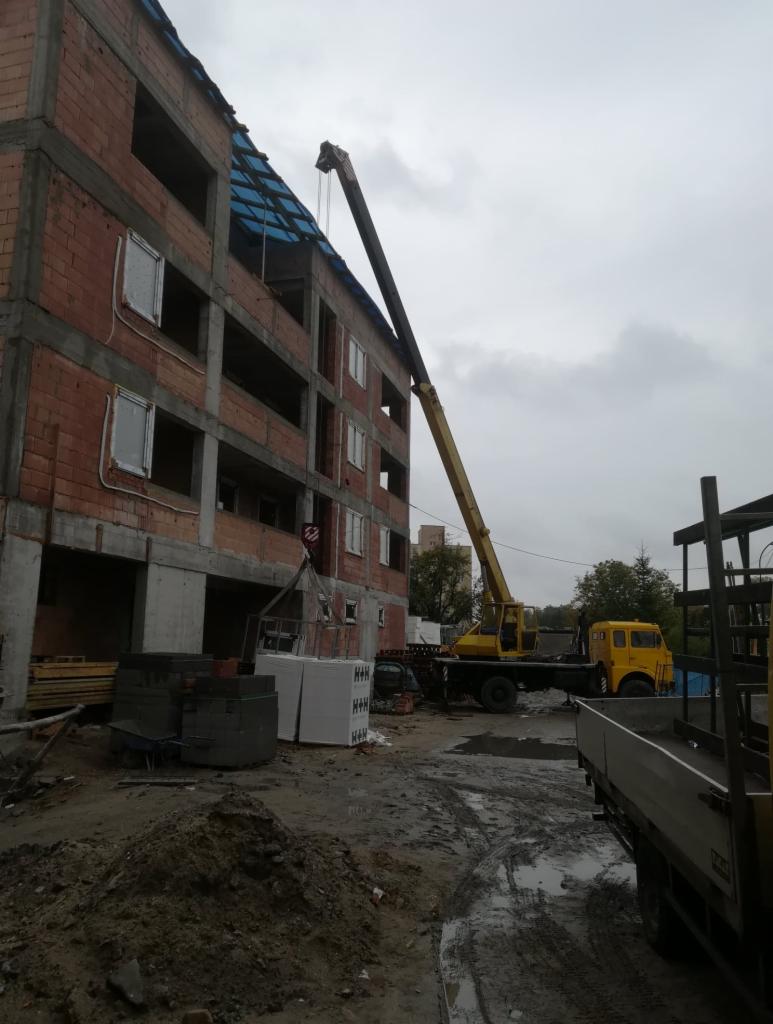 Żółty dźwig samochodowy podnosi materiały budowlane na budowę czteropiętrowego budynku z czerwonej cegły w pochmurny dzień.