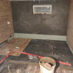 Łazienka w trakcie układania płytek podłogowych na ogrzewaniu podłogowym, z użyciem poziomicy i systemu poziomowania płytek, ściany częściowo wykończone panelami i płytkami imitującymi kamień.