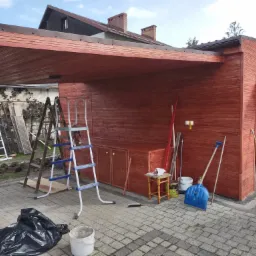 Drewniana konstrukcja z zadaszeniem, prawdopodobnie garaż lub wiata, świeżo po malowaniu, widoczne narzędzia malarskie, drabiny i worki na śmieci.