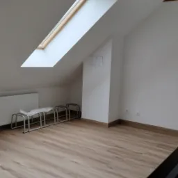 Poddasze po remoncie z drewnianymi panelami podłogowymi, oknem dachowym i grzejnikiem. Metalowe stojaki na rowery w rogu pomieszczenia.