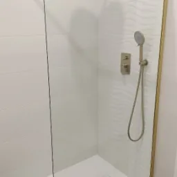 Kabina prysznicowa walk-in z brodzikiem, złotymi profilami i deszczownicą, na ścianach białe płytki z subtelnym falistym wzorem.