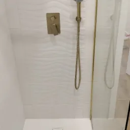 Kabina prysznicowa z białymi płytkami o falistej strukturze, złota armatura i szklane drzwi z złotym obramowaniem.