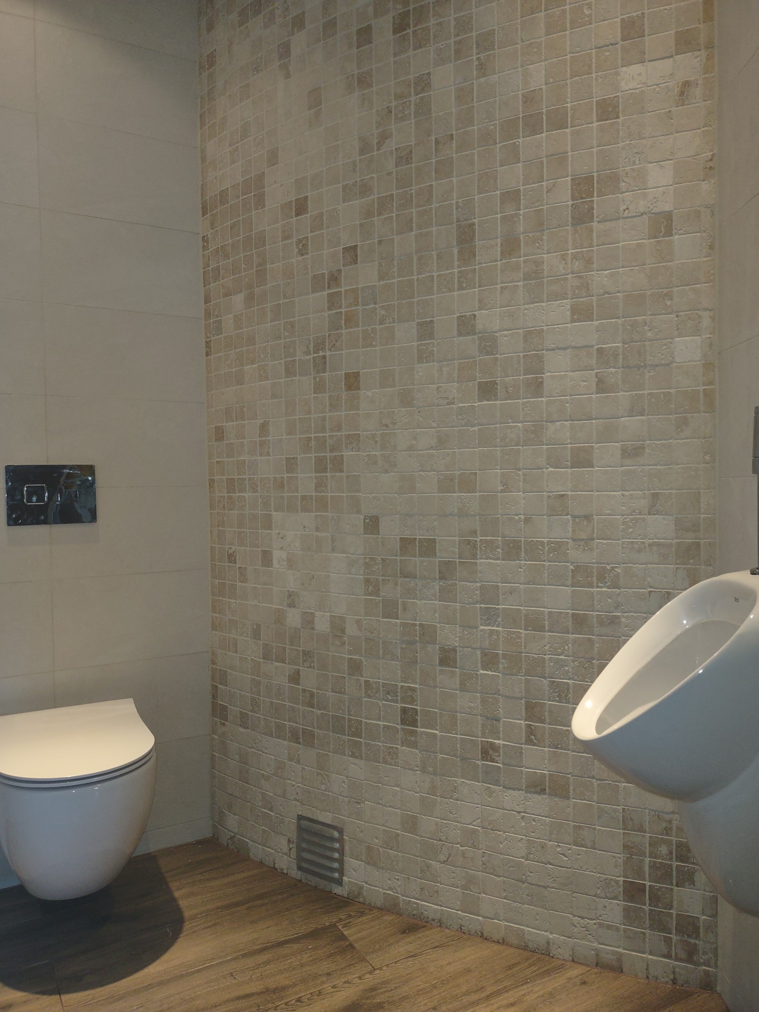 Nowoczesna łazienka z beżowymi mozaikowymi płytkami na ścianie, wiszącą toaletą i pisuaru, drewnopodobna podłoga i chromowana armatura.