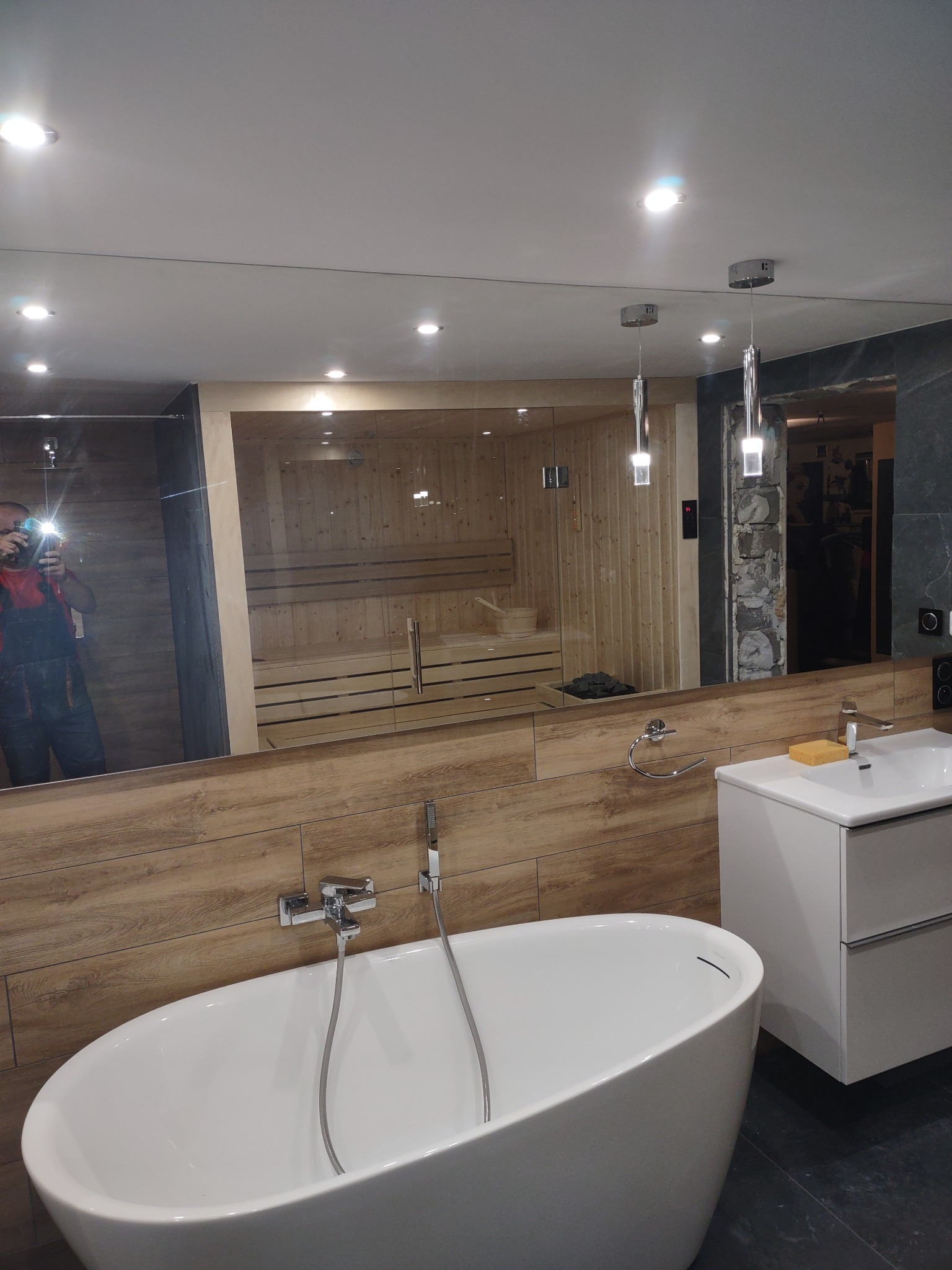 Nowoczesna łazienka z owalną wanną, umywalką nablatową i sauną widoczną w lustrze, oświetlenie punktowe i wiszące lampy, ściany wykończone płytkami imitującymi drewno i ciemny kamień.