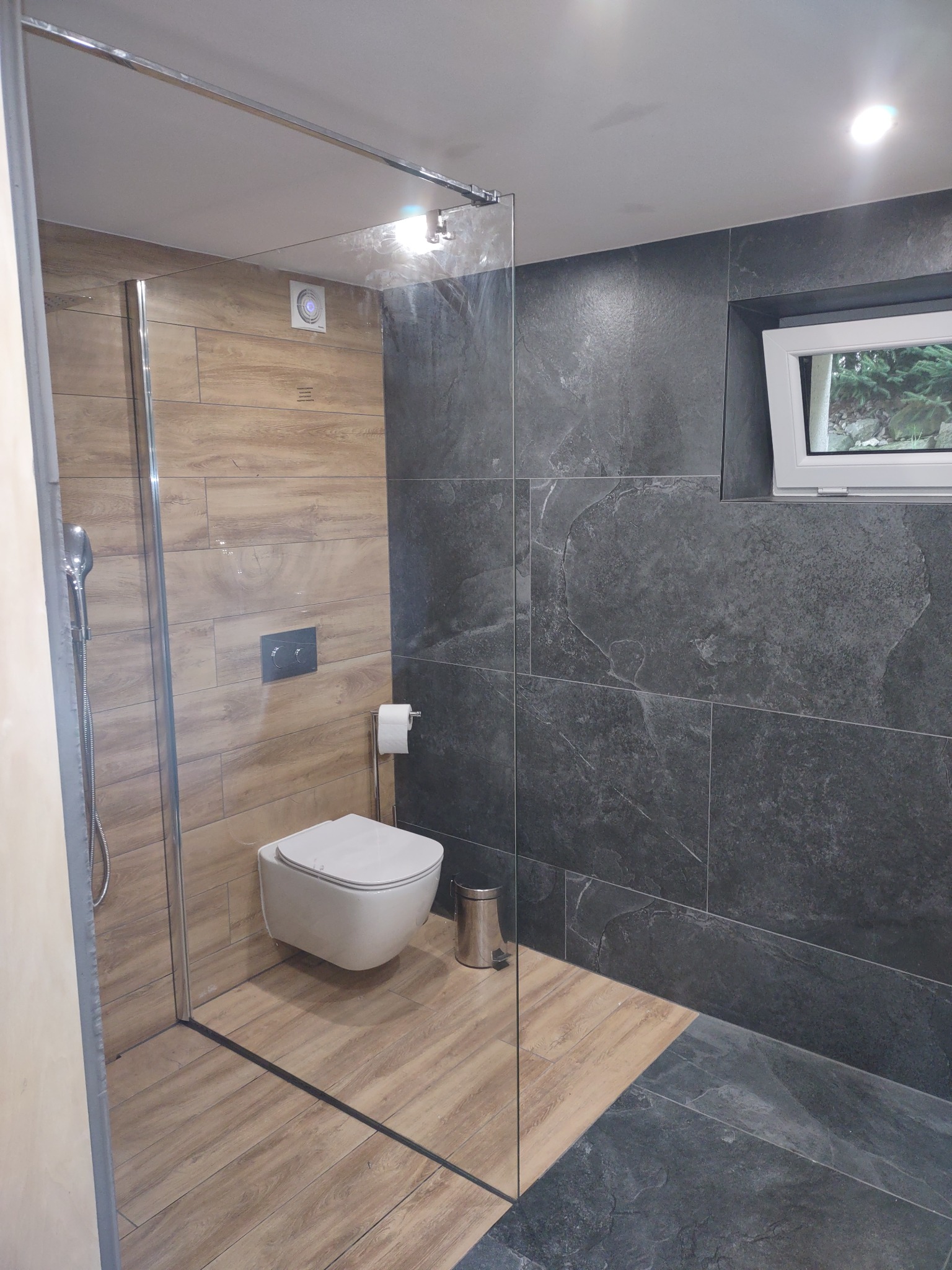Nowoczesna łazienka z kabiną prysznicową typu walk-in, wiszącą toaletą i ścianami wyłożonymi płytkami w odcieniach drewna i szarości, z widocznym oknem w tle.