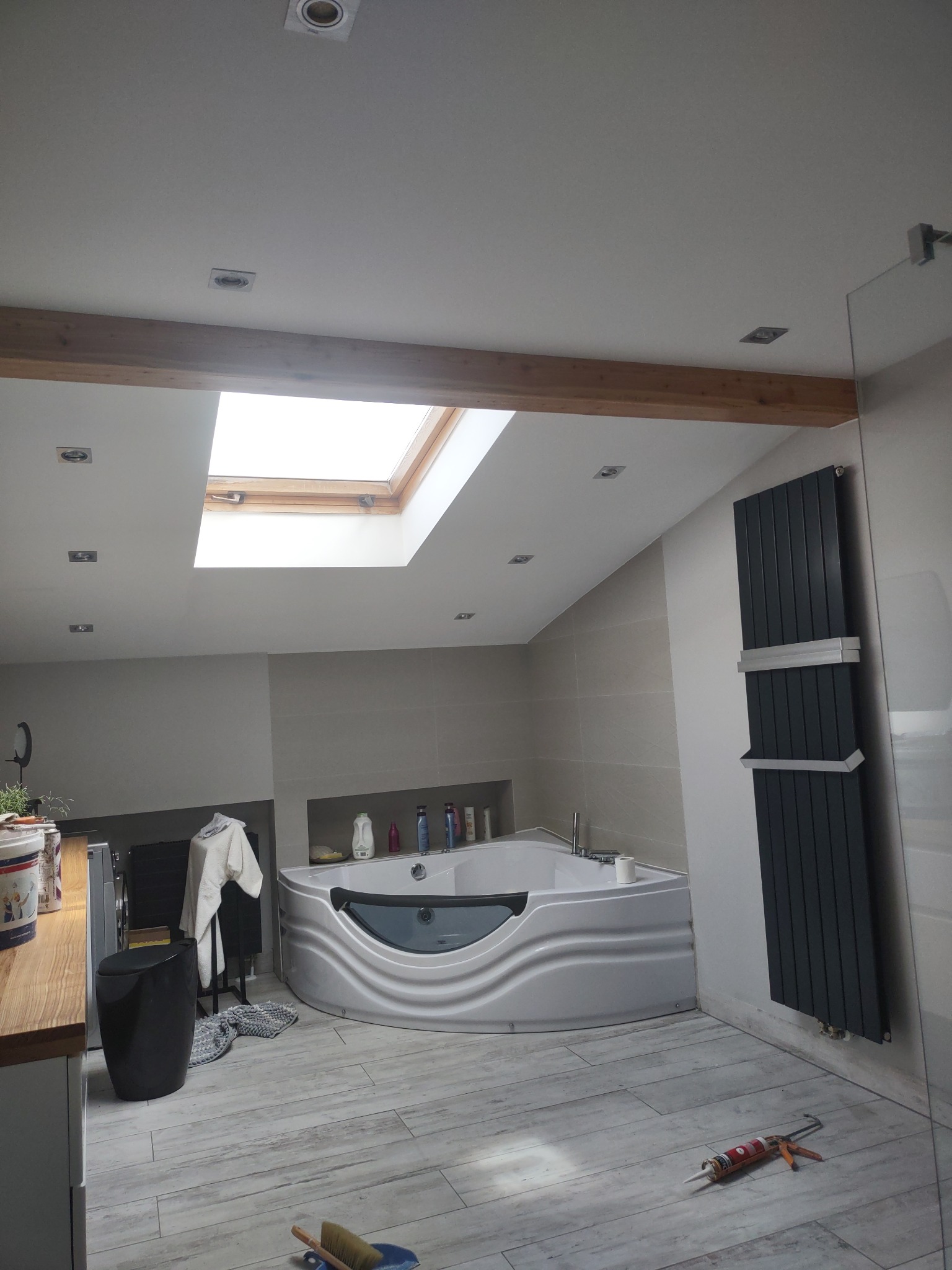 Przestronna łazienka na poddaszu z oknem dachowym, narożną wanną z hydromasażem i grzejnikiem drabinkowym w kolorze antracytowym. Na podłodze płytki imitujące drewno, widoczne narzędzia i silikon...