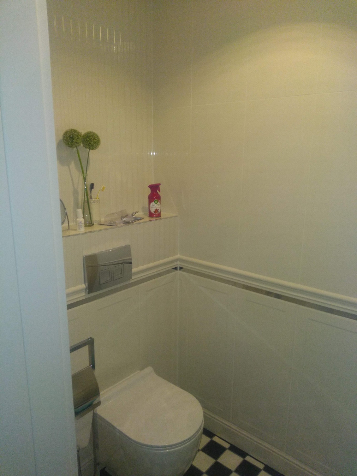 Wnętrze toalety po remoncie: białe płytki ścienne z szarą listwą, czarno-biała szachownica na podłodze, wiszące WC z chromowanym przyciskiem spłukiwania, dekoracje na półce.