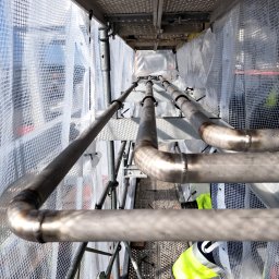 Firma Usługowa Jacek Chojecki - Spojrzenie z góry na spawane rury na rusztowaniu, zabezpieczone siatką ochronną. Widoczne złącza spawane i fragment stroju roboczego.