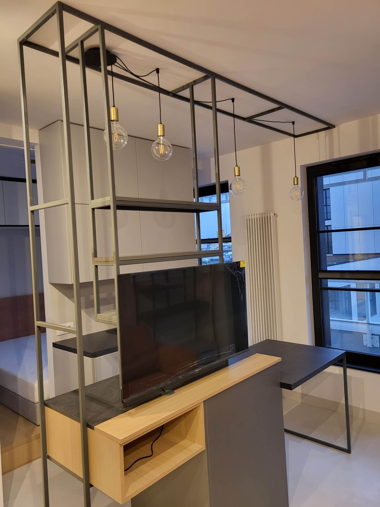 Metalowa konstrukcja regału z oświetleniem w nowoczesnym wnętrzu. Widoczne spawane elementy, półki, lampy z żarówkami Edisona i biurko z metalowymi nogami.