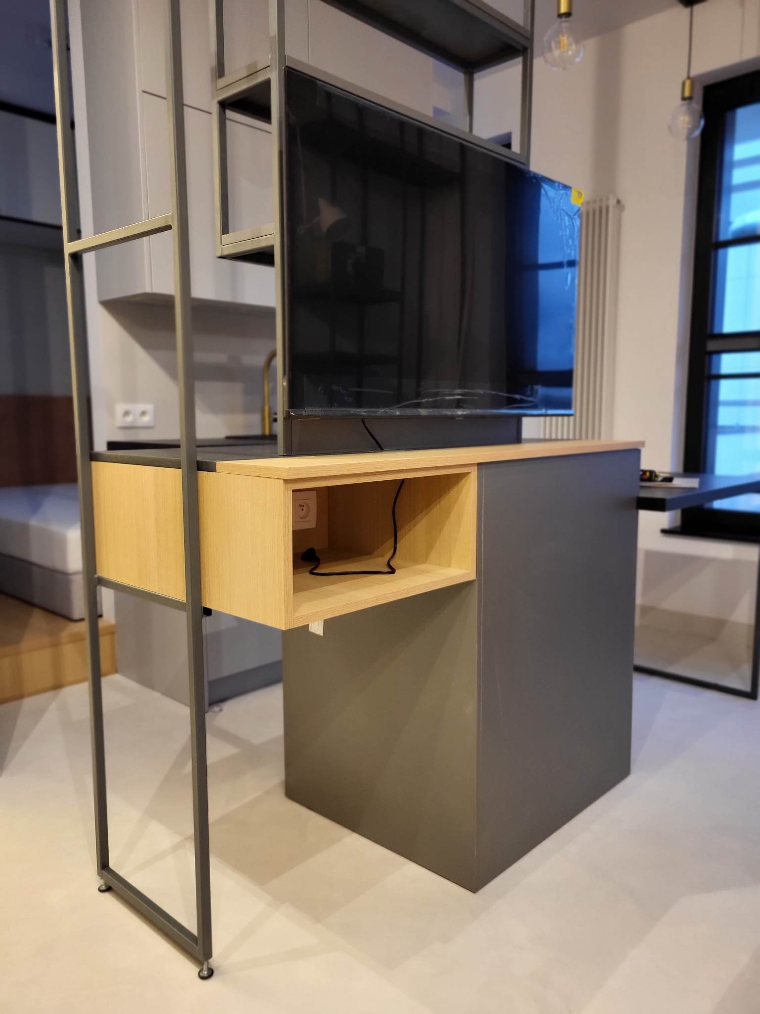 Nowoczesny regał z metalową ramą i drewnianymi elementami, wbudowany w ścianę, z półkami i miejscem na telewizor. Minimalistyczny design, szara kolorystyka.