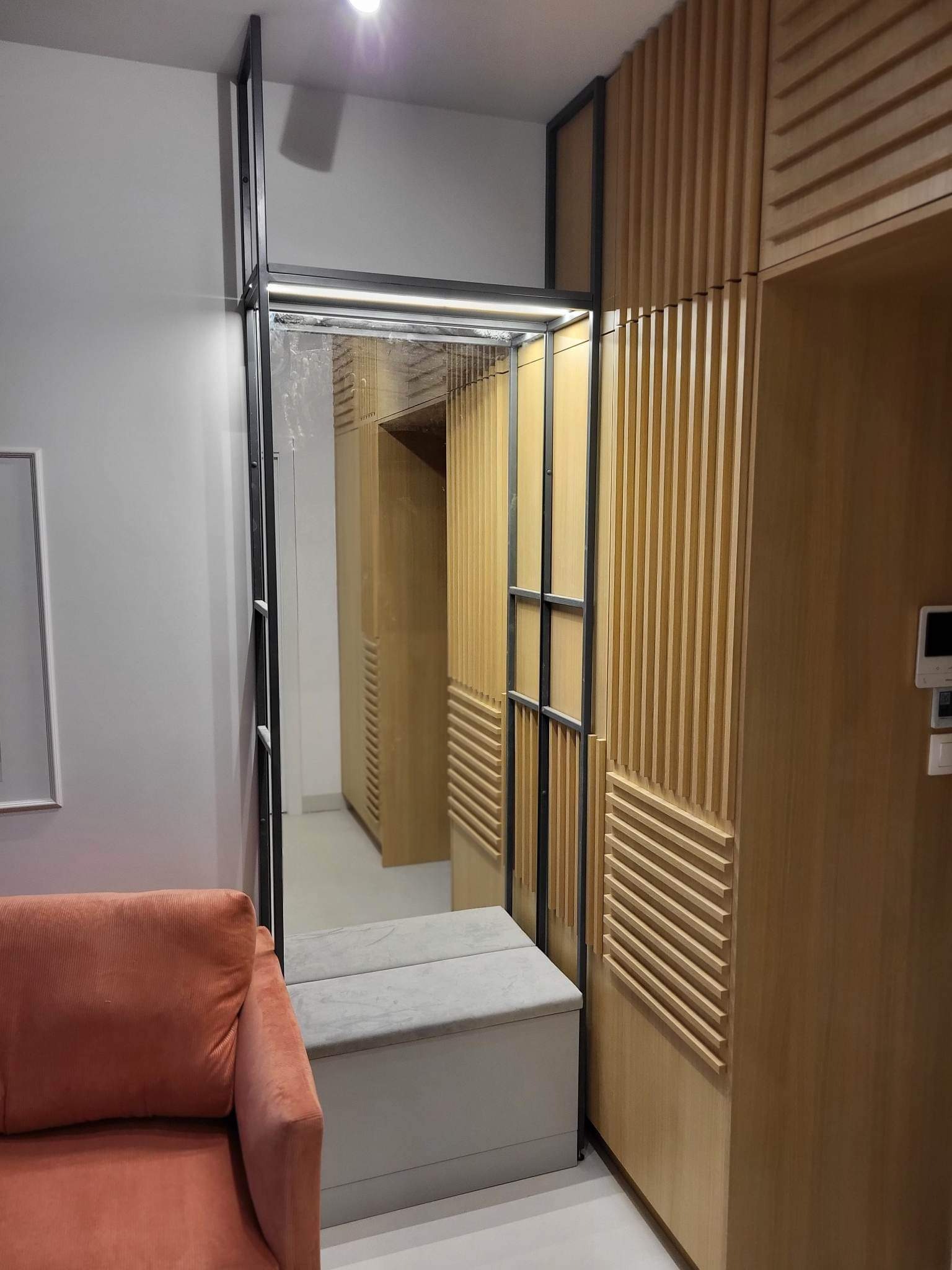 Nowoczesna zabudowa przedpokoju z lustrem w metalowej ramie, drewnianymi panelami ściennymi i siedziskiem. Minimalistyczny design, ciepłe oświetlenie.