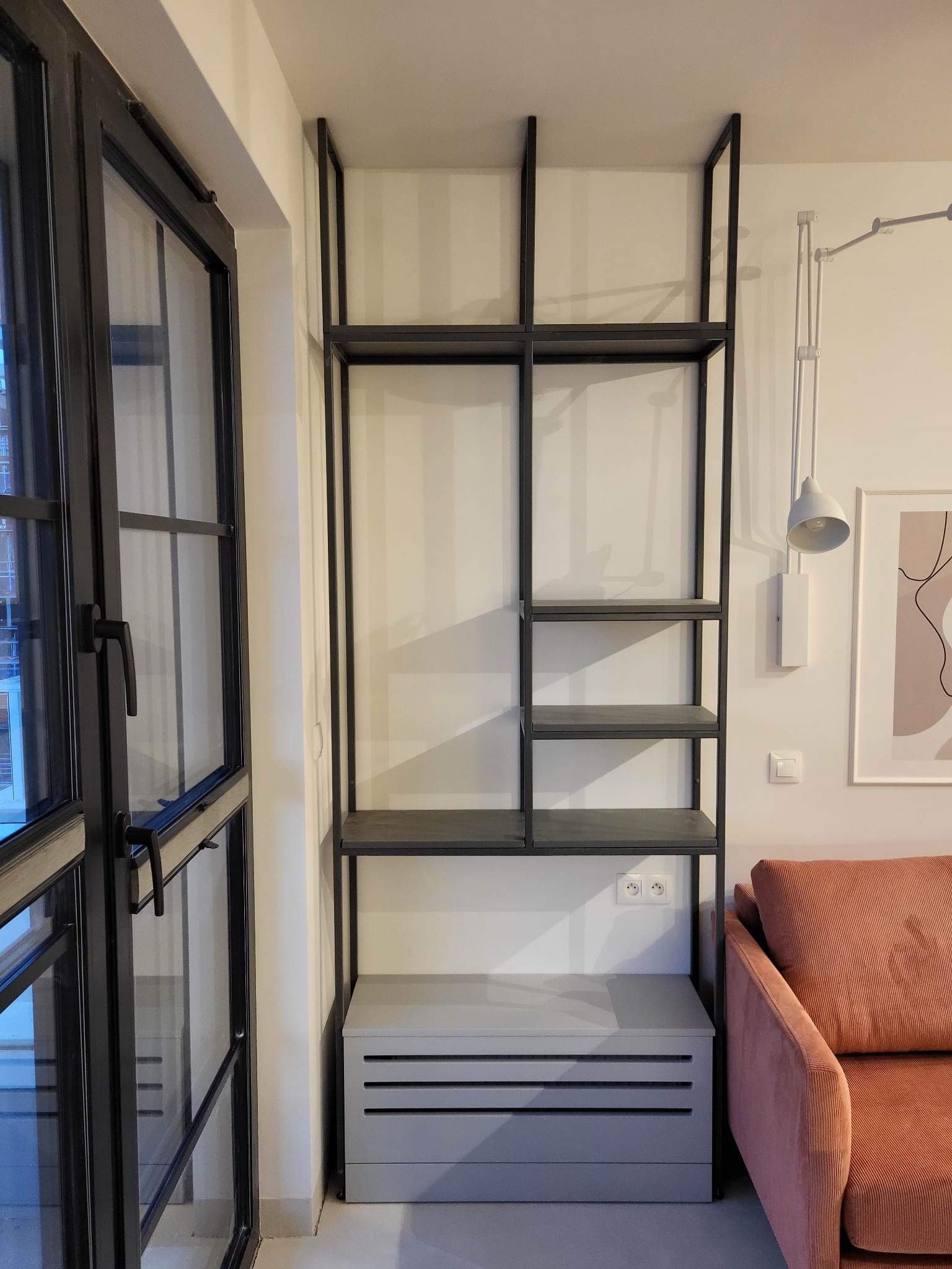 Metalowy regał w stylu loft z szarymi półkami, obok okno z czarną ramą i różowa sofa. Minimalistyczne wnętrze z lampą ścienną i obrazem.