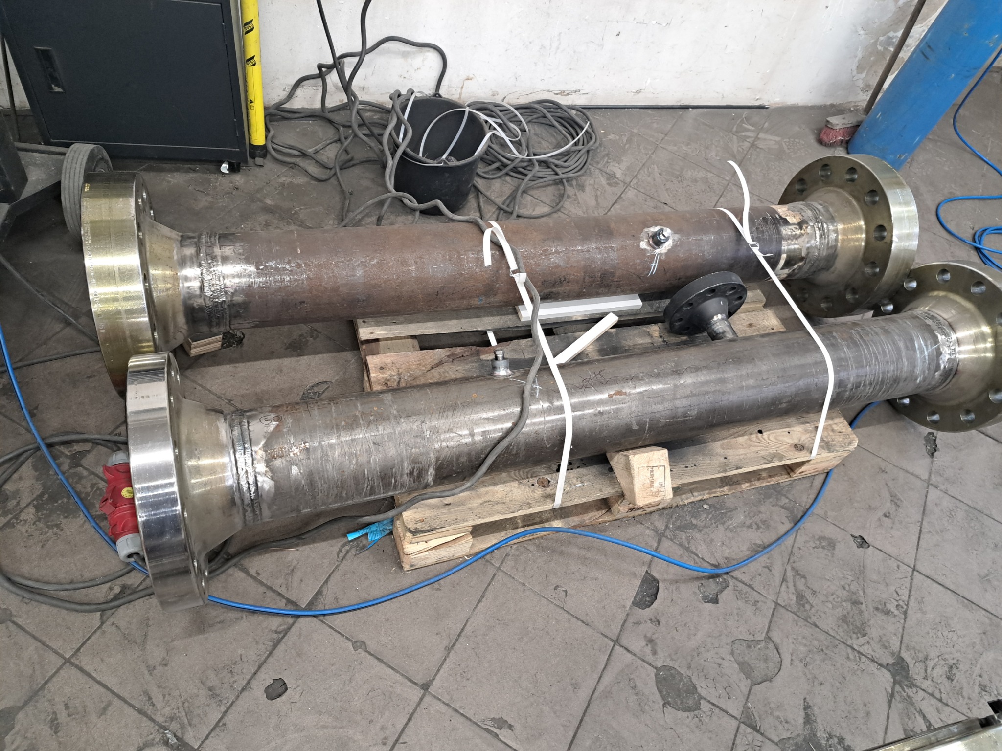 Dwie spawane rury z kołnierzami na palecie, widoczne spawy, kable, w tle sprzęt spawalniczy i wiadro z kablami. Szara podłoga z widocznymi śladami użytkowania.