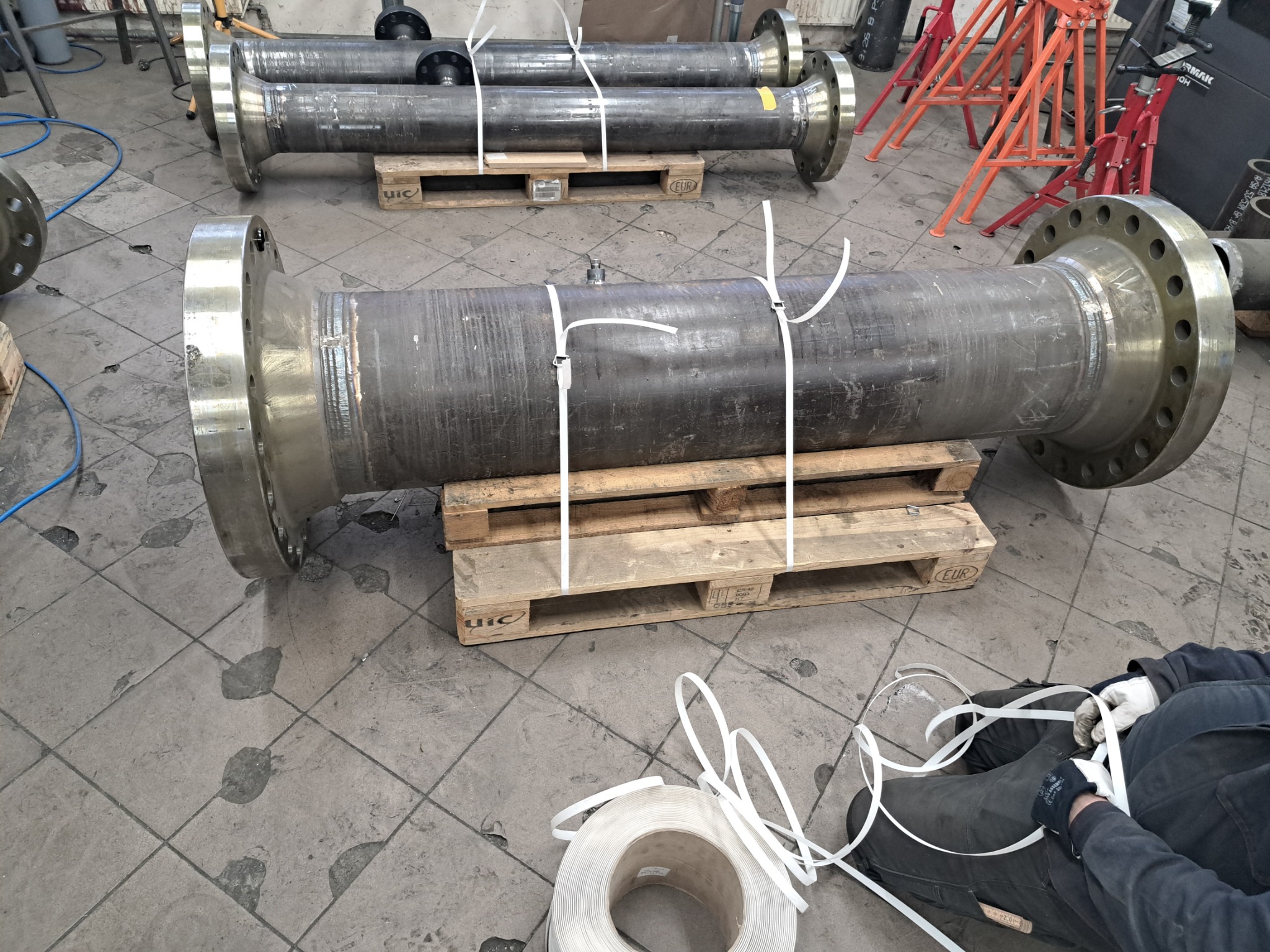 Spawane rury z kołnierzami na paletach, zabezpieczone taśmą. Widoczne ślady spawania i obróbki metalu. Praca w warsztacie.