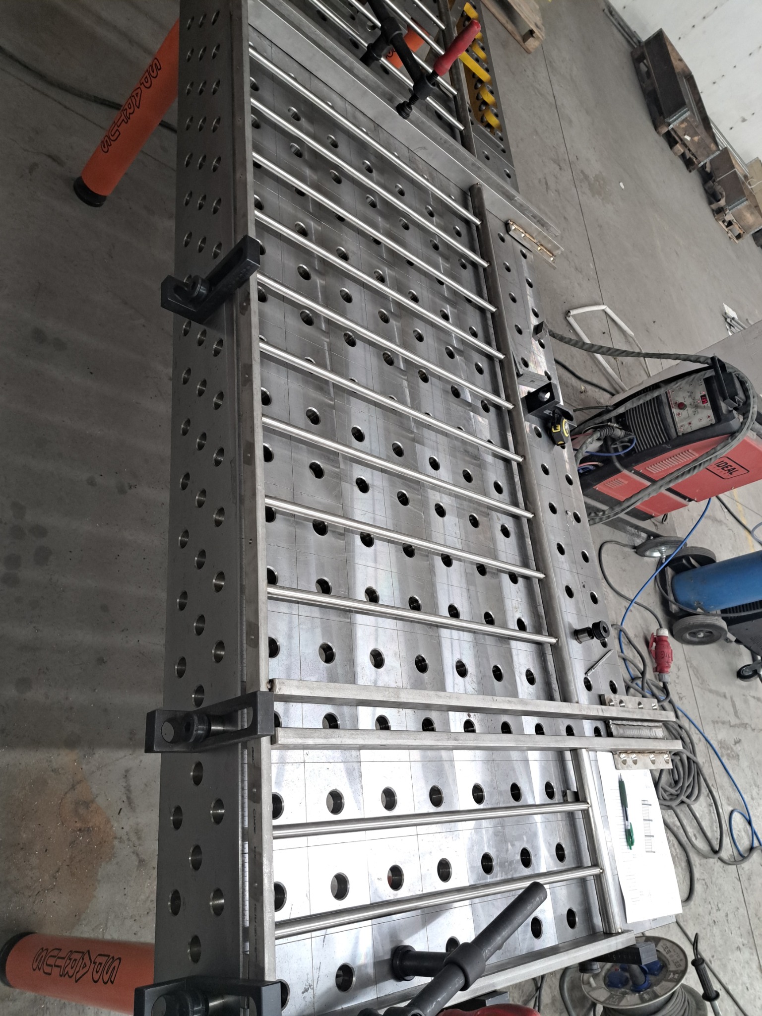 Stół spawalniczy z perforacją i zamocowanymi elementami, widoczny sprzęt spawalniczy w tle. Metalowa konstrukcja z licznymi otworami.