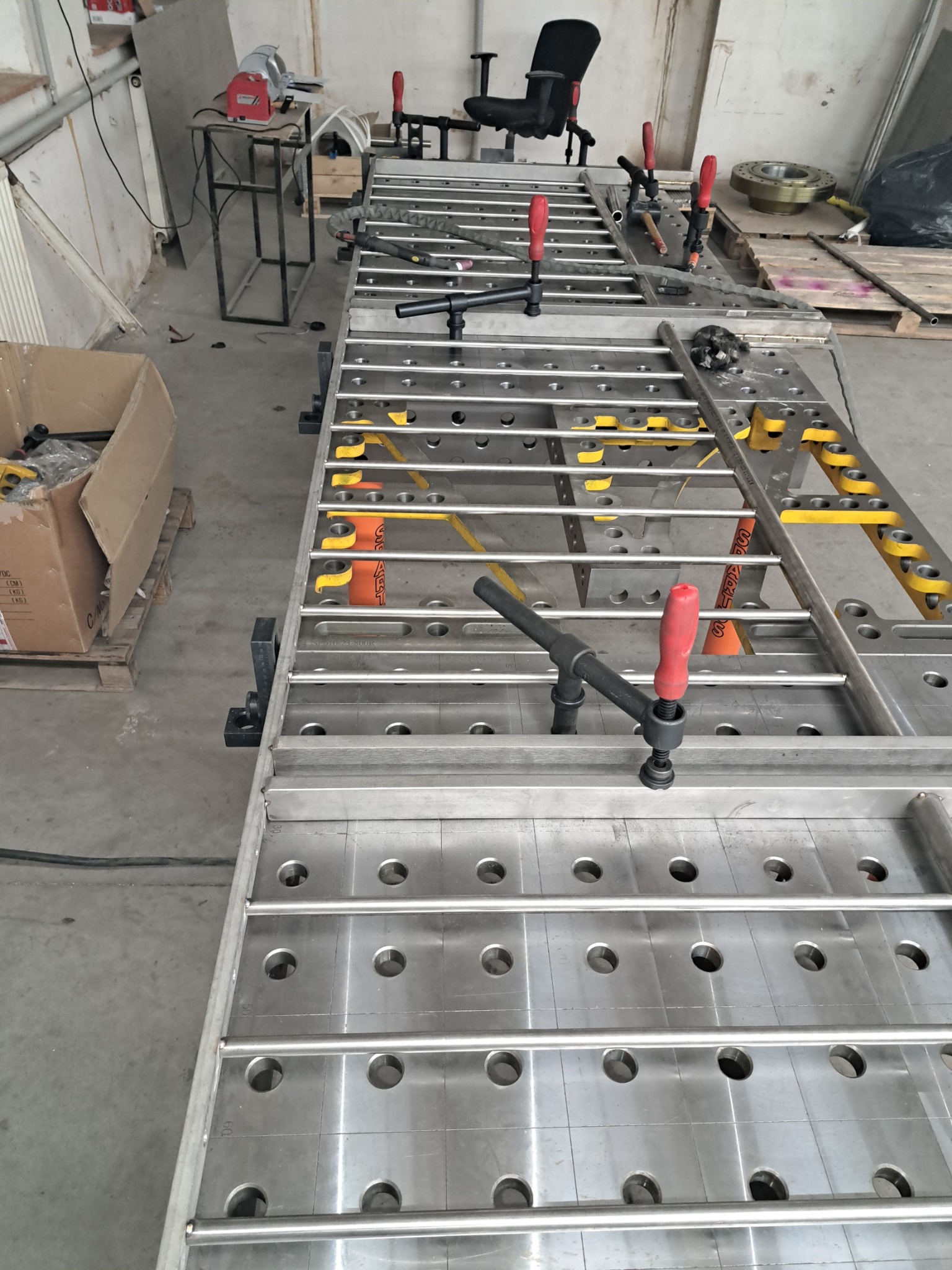 Stół spawalniczy z elementami mocującymi, widoczne otwory i pręty, narzędzia na stole, w tle warsztat, przygotowanie do pracy, kątowe ujęcie, oświetlenie warsztatowe.